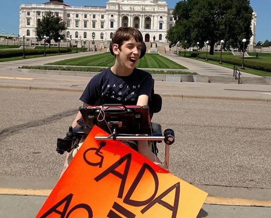Justin in wheelchair holding ADA = Access sign in front of MN State Capitol