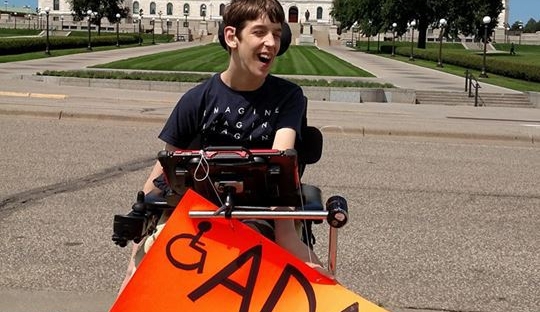 Justin in wheelchair holding ADA = Access sign in front of MN State Capitol