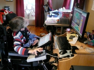 Justin in wheelchair with hand on joystick mouse looking at computer and communication device