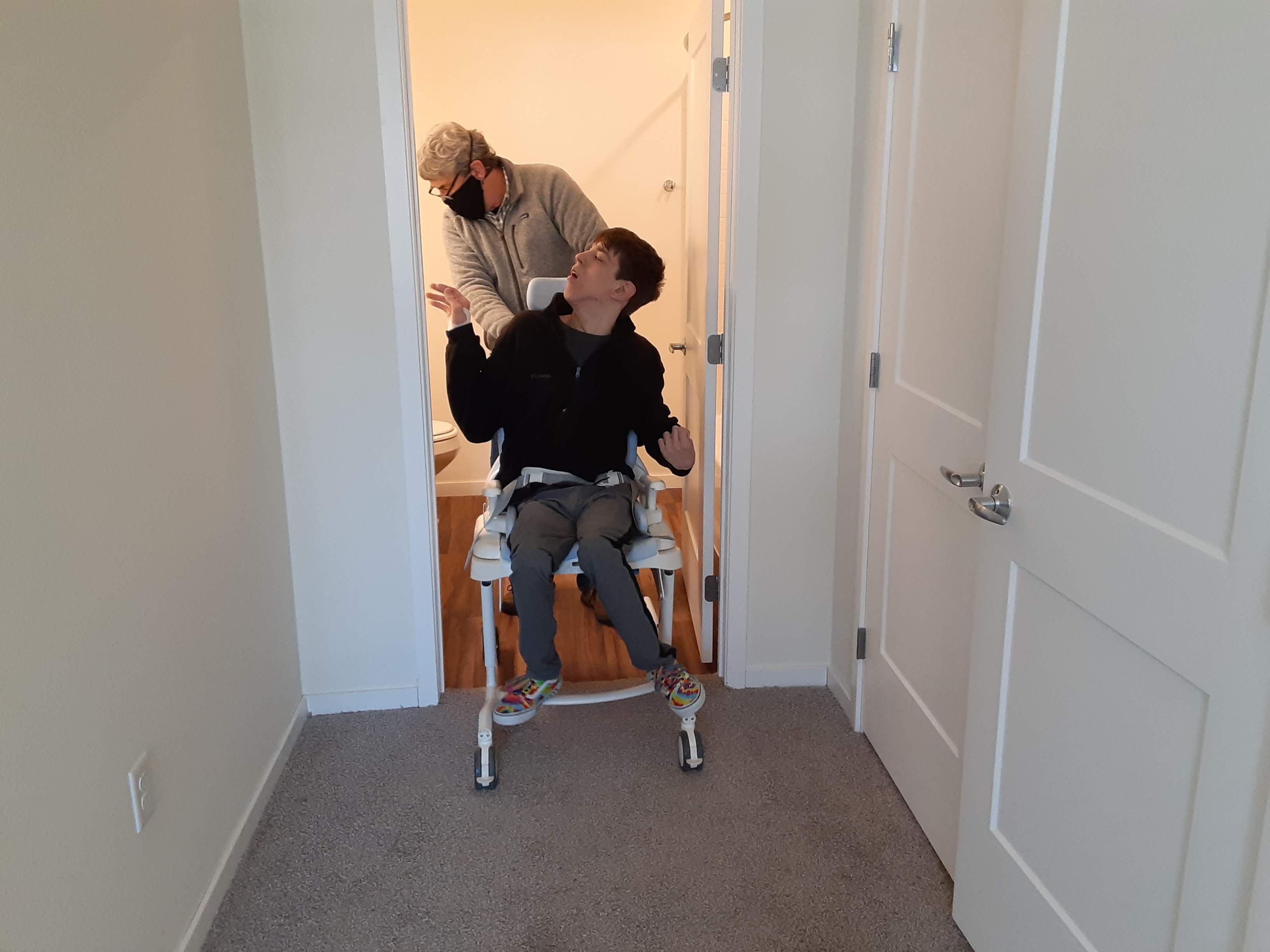 Justin in custom shower chair rolling through doorway into bathroom, dad behind him
