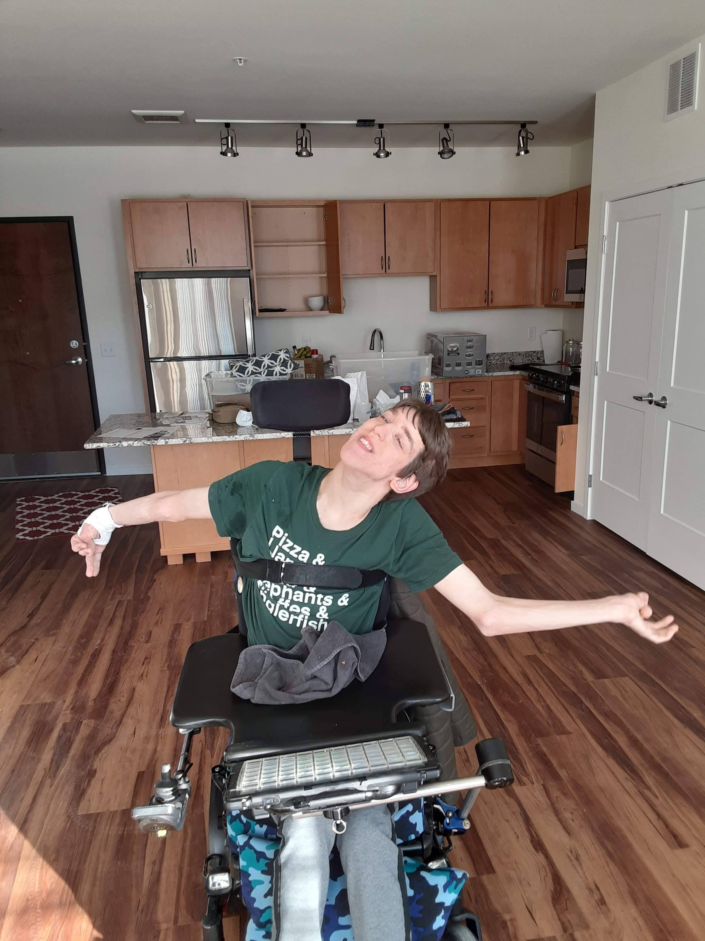 Justin in power wheelchair, smiling with arms outstretched in new empty apartment, kitchen behind him