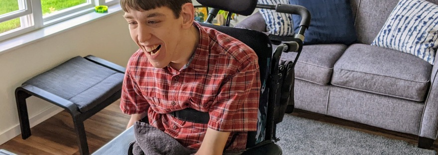 Justin, young smiling man seated in wheelchair in front of adapted keyboard, in living room