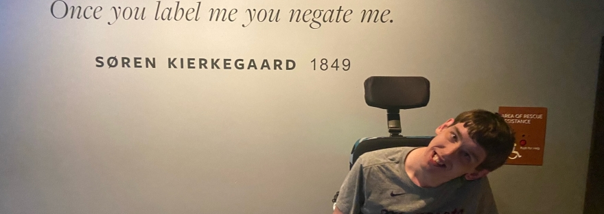 Young man in wheelchair in front of quote painted on wall by Soren Kierkegaard, 1849, "Once you label me you negate me."