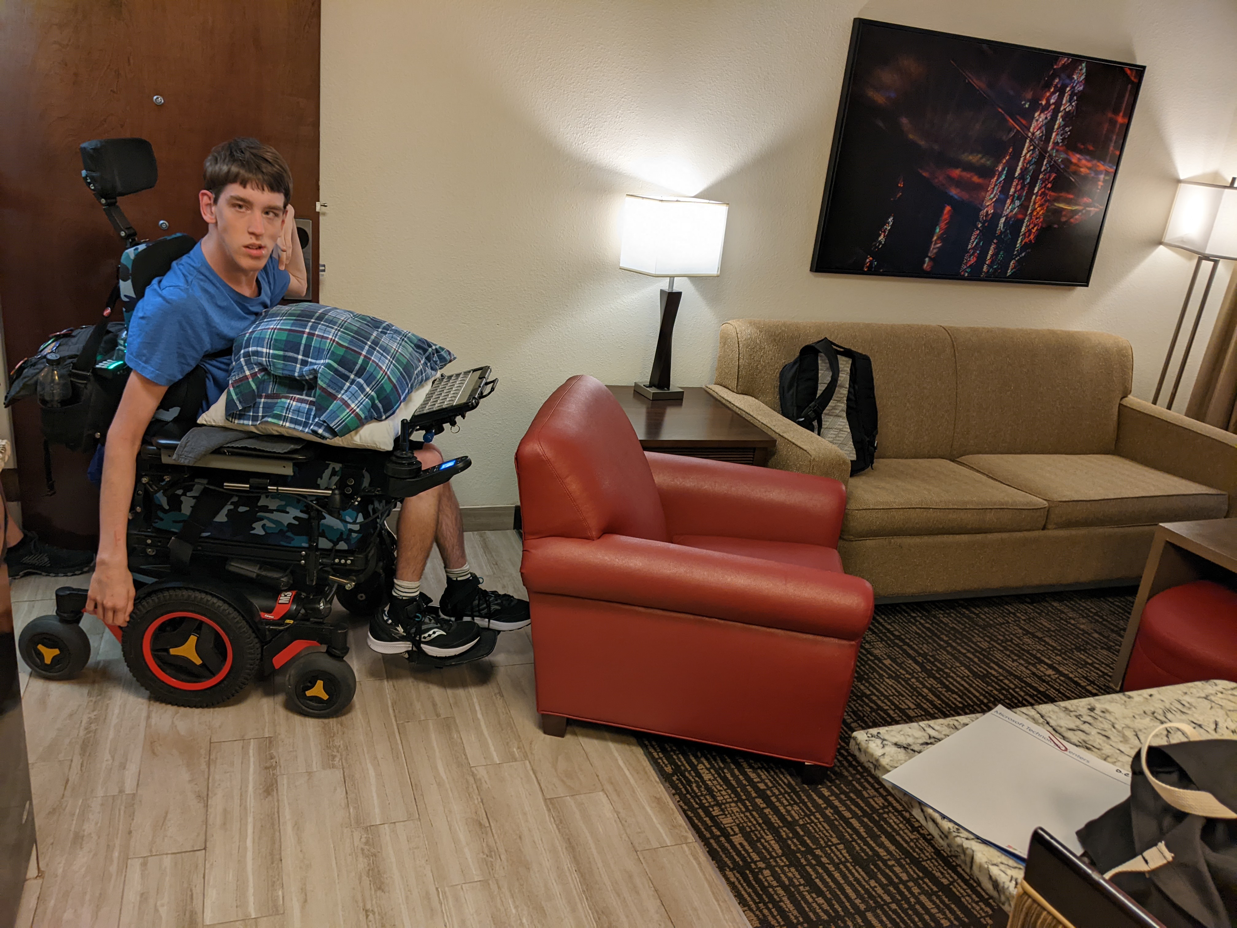 Young man in power wheelchair holding a pillow in doorway, big easy chair in front of him, couch
