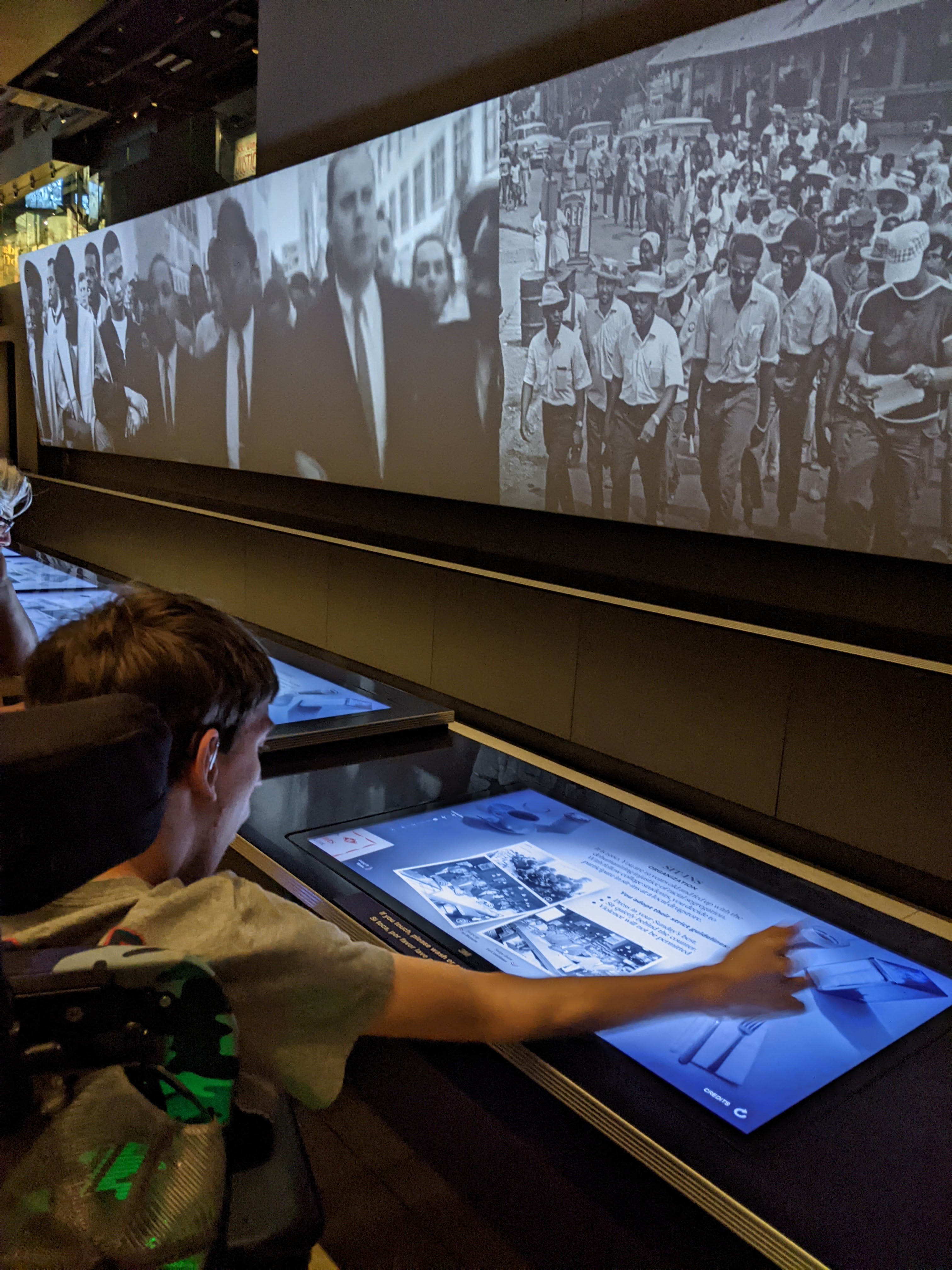 Young man in wheelchair looking at interactive monitor at exhibit with images on large screen from 1960s civil rights era