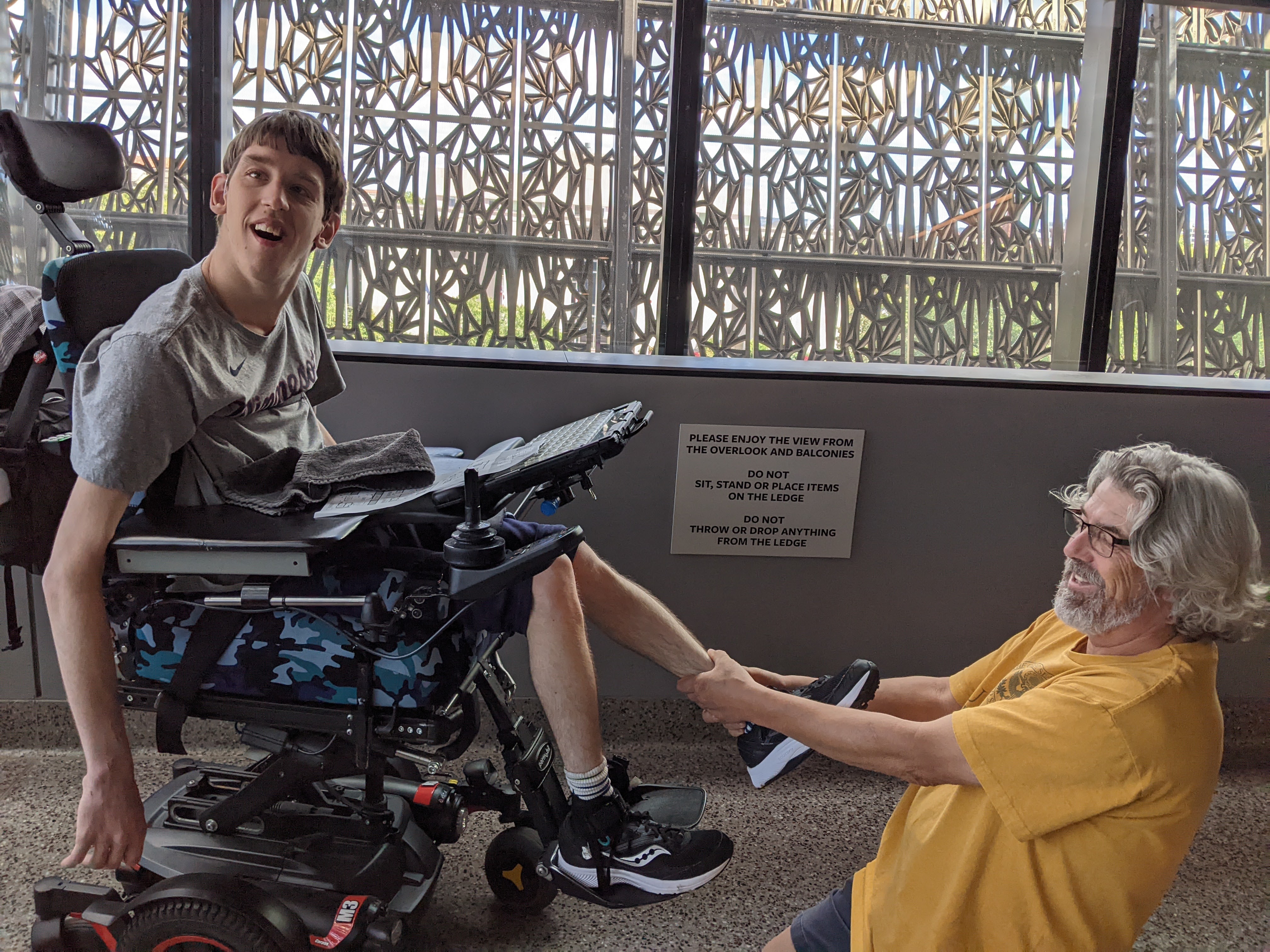 Young man in raised up in power wheelchair with his dad stretching young man's leg