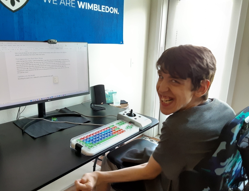 Justin, young man, seated in wheelchair in front of adapted computer keyboard and joystick mouse, smiling at camera, Word document open on computer monitor, We are Wimbledon flag above monitor