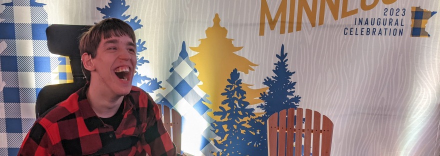 Justin, young man laughing, wearing red and black houndstooth plaid shirt, seated in power wheelchair, in front of One Minnesota 2023 Inaugural Celebration banner with yellow and blue pine trees