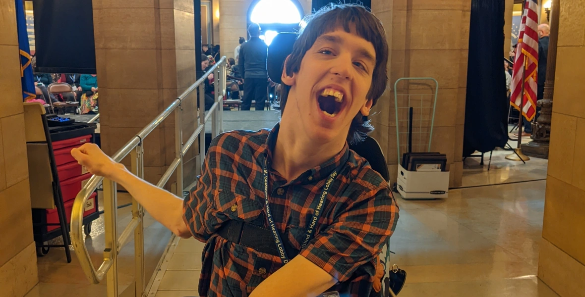 Justin, young man in his 20s seated in power wheelchair with big smile, ramp behind to a stage in the MN Capitol Rotunda