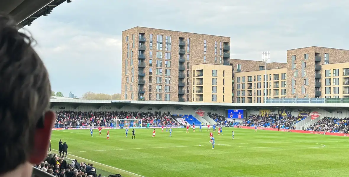 back of Justin's head looking at AFC Wimbledon team playing on pitch, buildings and crowded stadium