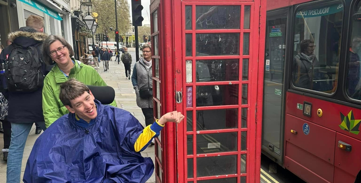 Justin, young man smiling in wheelchair wearing blue rain poncho and his mom standing behind, red telephone booth and red London doubledecker bus next to them, British flag hanging across the street, people walking on sidewalk