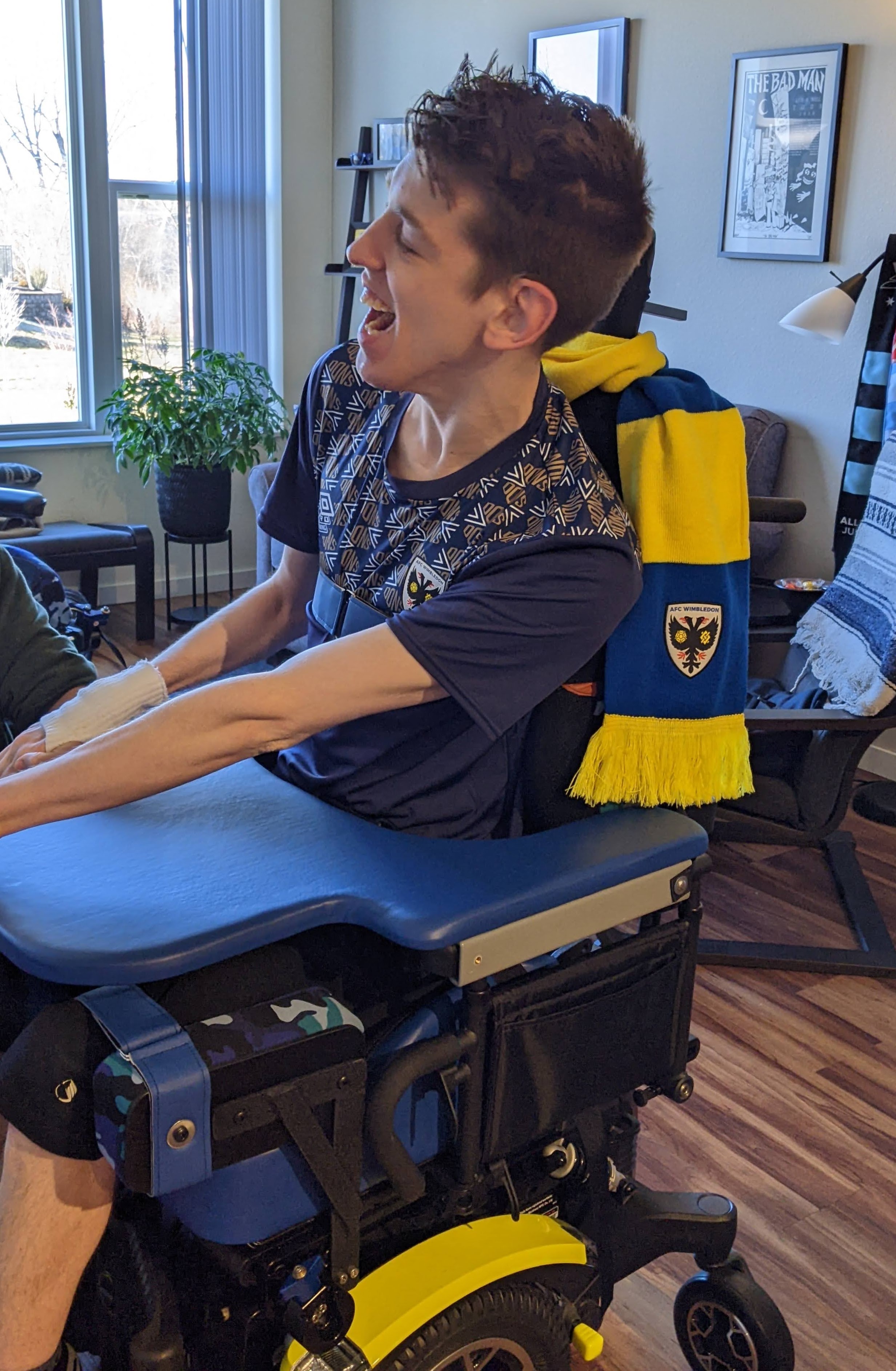 Justin, a smiling young man in his 20s, sitting in blue and yellow power wheelchair, blue and yellow AFC Wimbledon scarf wrapped around headrest