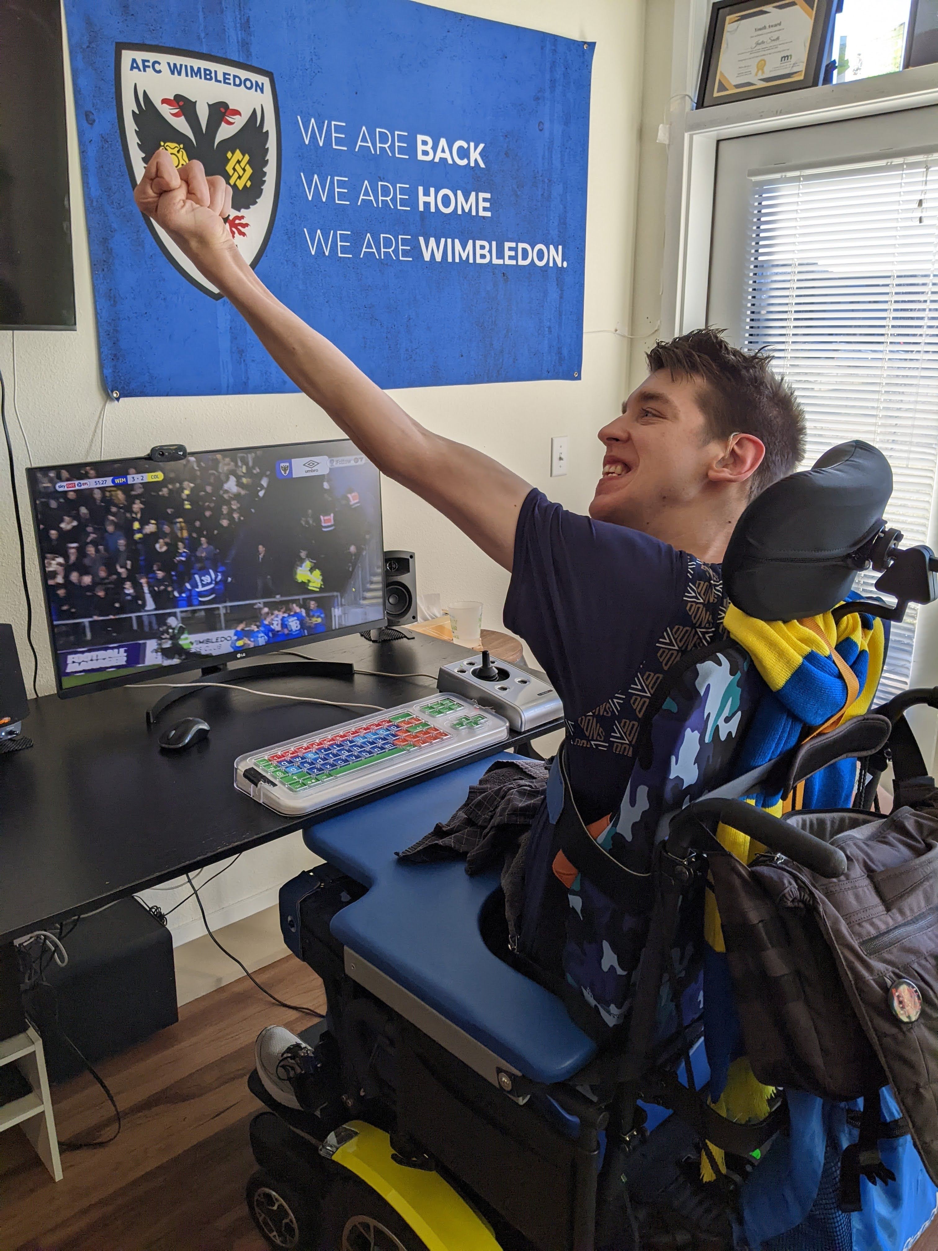 Justin, a smiling young man in his 20s holding up arm in celebratory cheer as he watches soccer on computer, sitting in blue and yellow power wheelchair, blue and yellow AFC Wimbledon scarf wrapped around headrest, there is an AFC Wimbledon flag hanging on the wall.