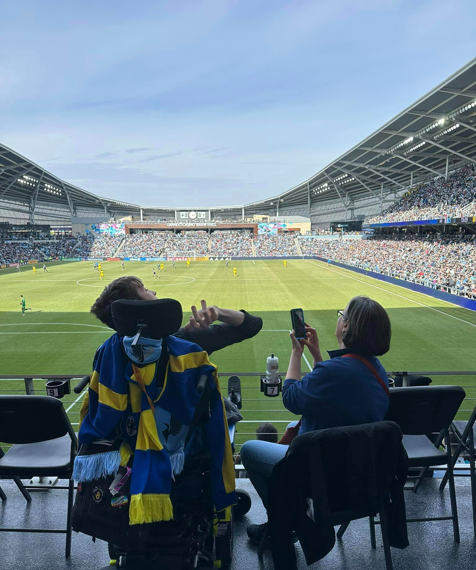 Back of Justin in his power wheelchair with soccer scarves hanging from the back, his mom sitting next to him taking a picture with cell phone overlooking MN United pitch and players