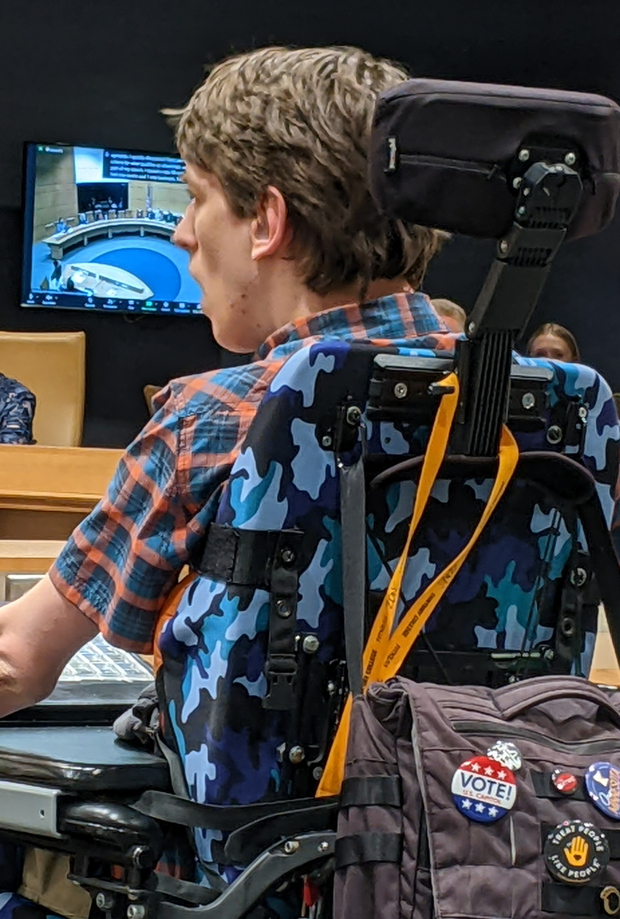 Back of Justin seated in his wheelchair, buttons on the back of his chair say Vote and Treat People Like People, monitor shows senate committee room