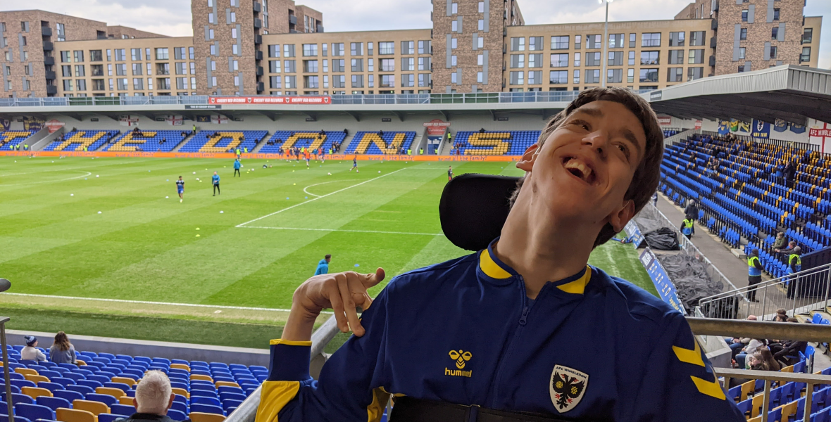 Justin smiling wearing a blue and yellow AFC Wimbledon jacket, Wimbledon football stadium behind with players warming up on the pitch